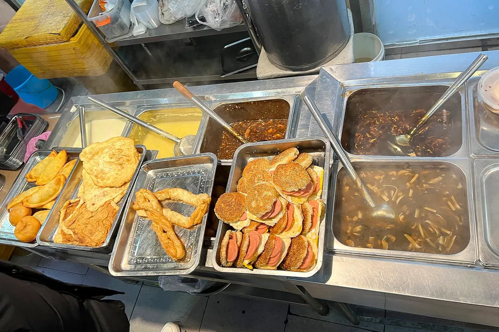 A Chinese breakfast spread of youtiao, soy milk, buns and savoury soups in warming trays