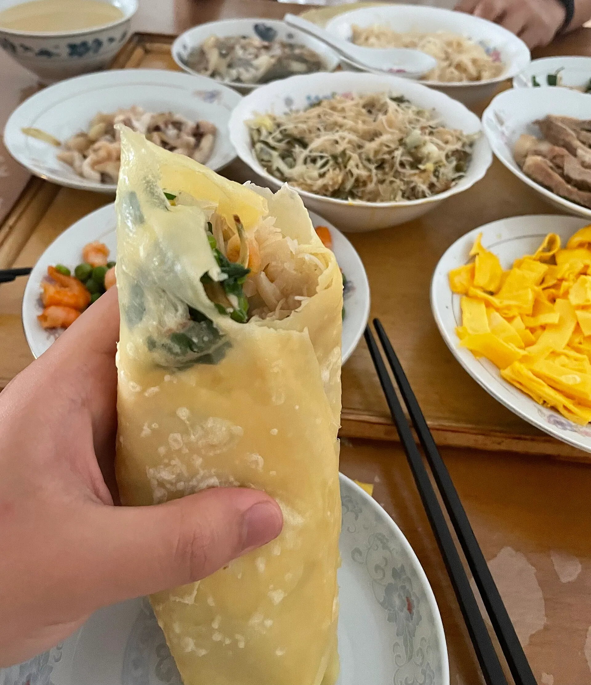 A hand holding a freshly wrapped shibingtong, with bowls of fillings on the table behind