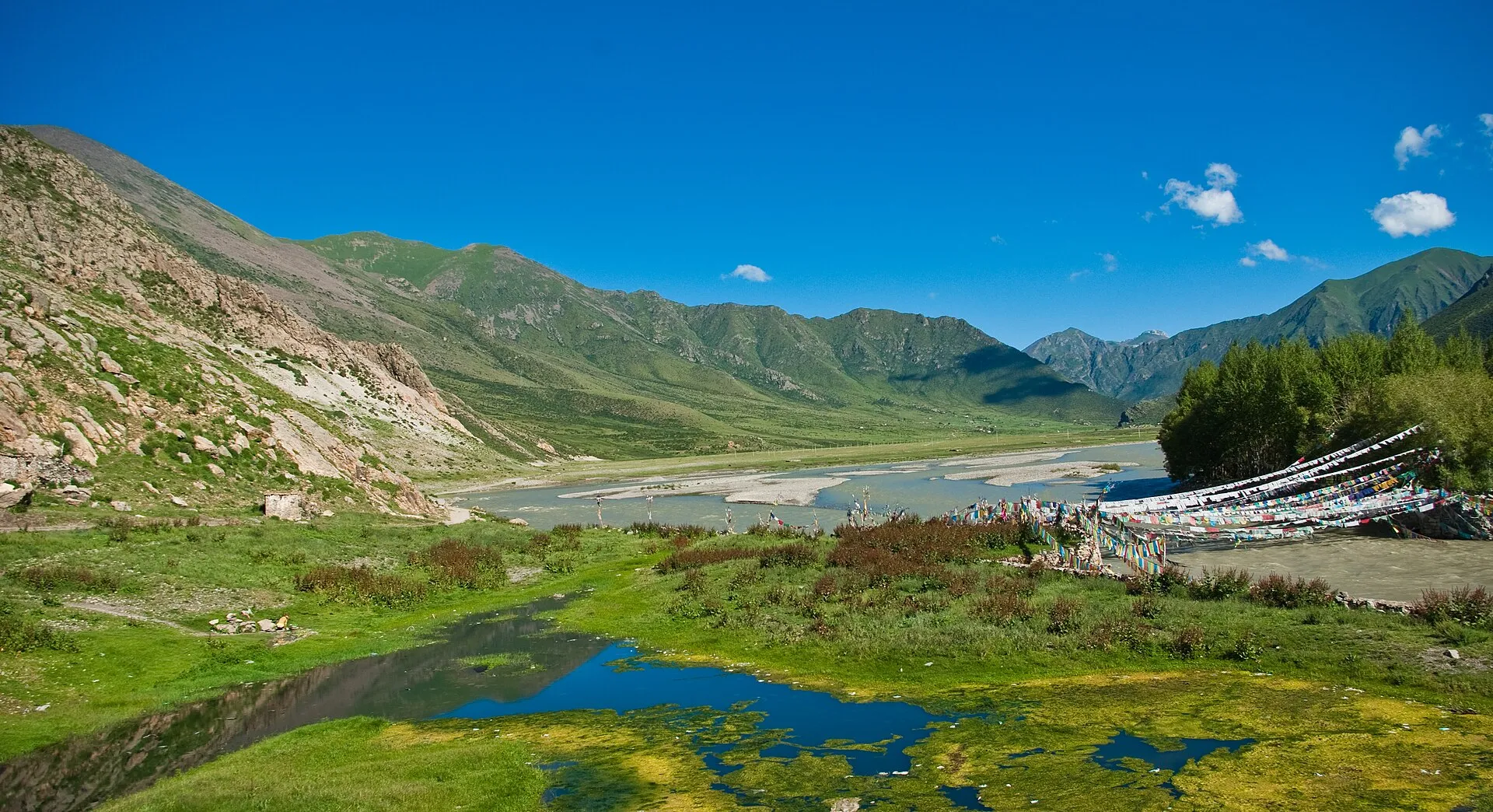 A Tibetan highland valley with prayer flags strung above a river, ringed by green mountains under a deep blue sky