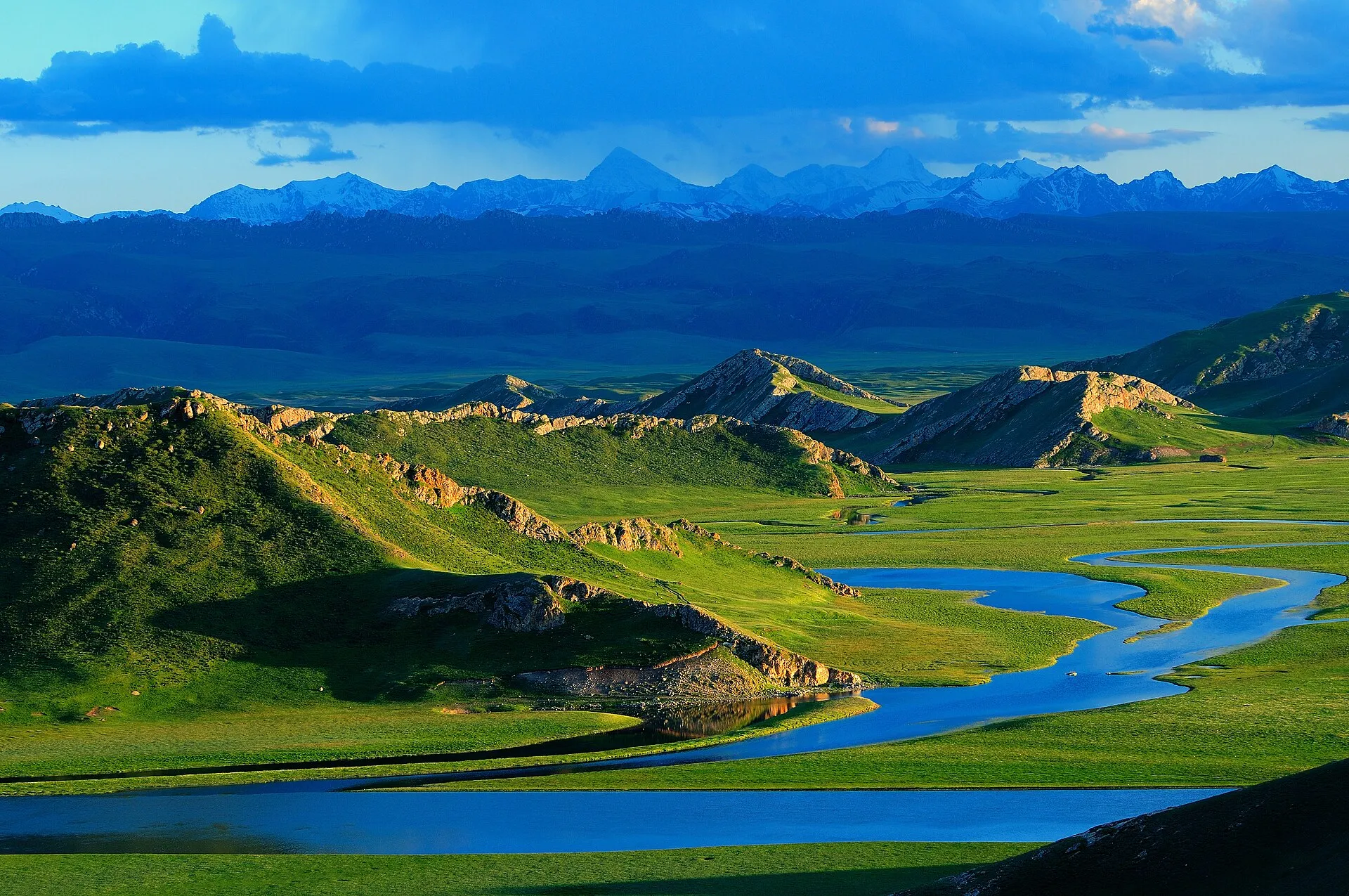 Snow-capped Tianshan peaks behind green grassland with a clear blue river winding through it under a blue sky, at Bayanbulak