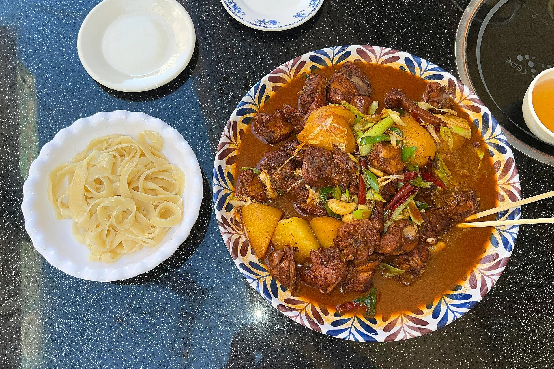 A bowl of Xinjiang big-plate chicken (大盘鸡) — chicken and potato in a rich, spicy sauce — served with a side of wide hand-pulled noodles