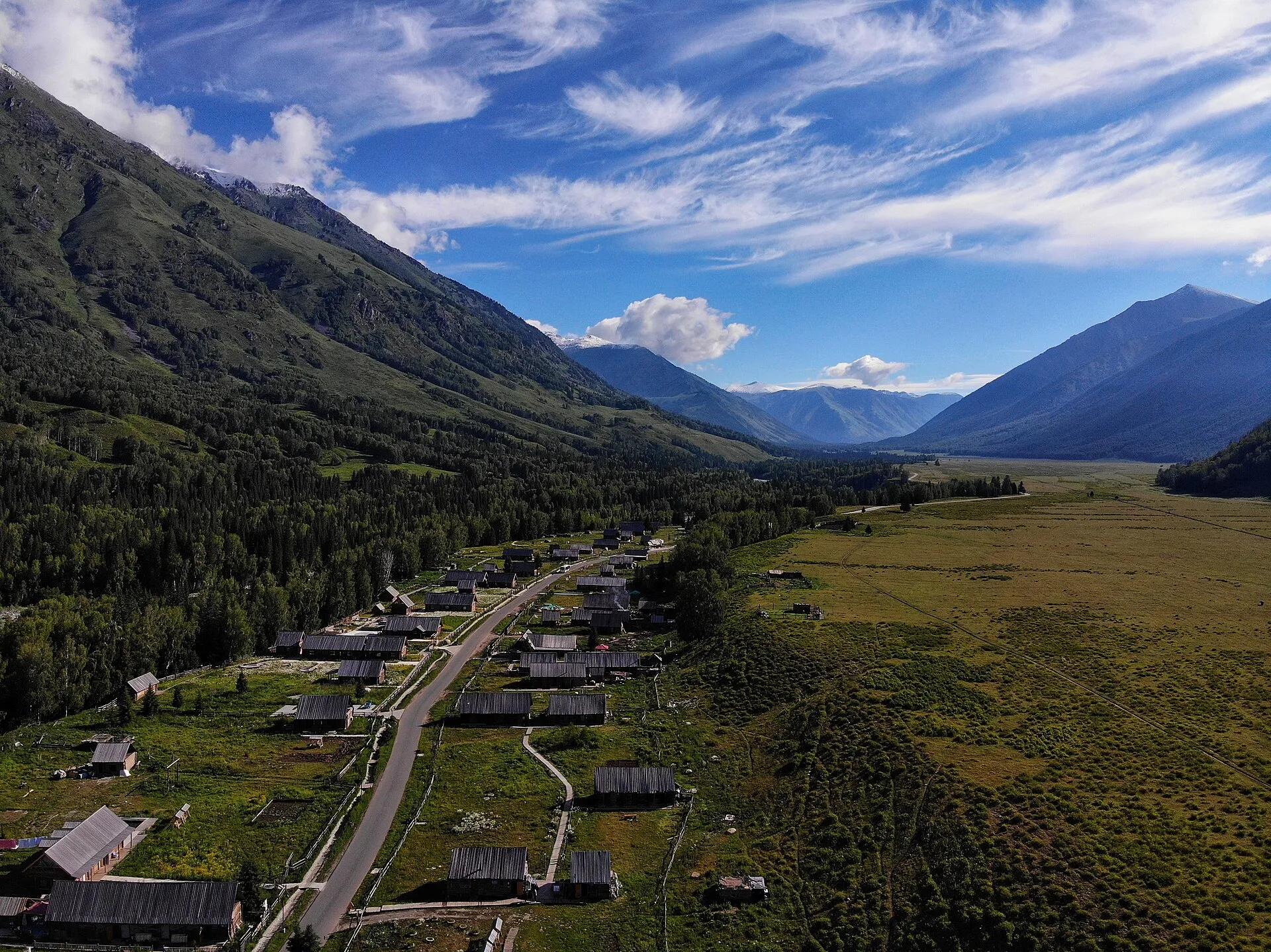The log cabins of Hemu village spread along a green valley floor, ringed by forested mountains