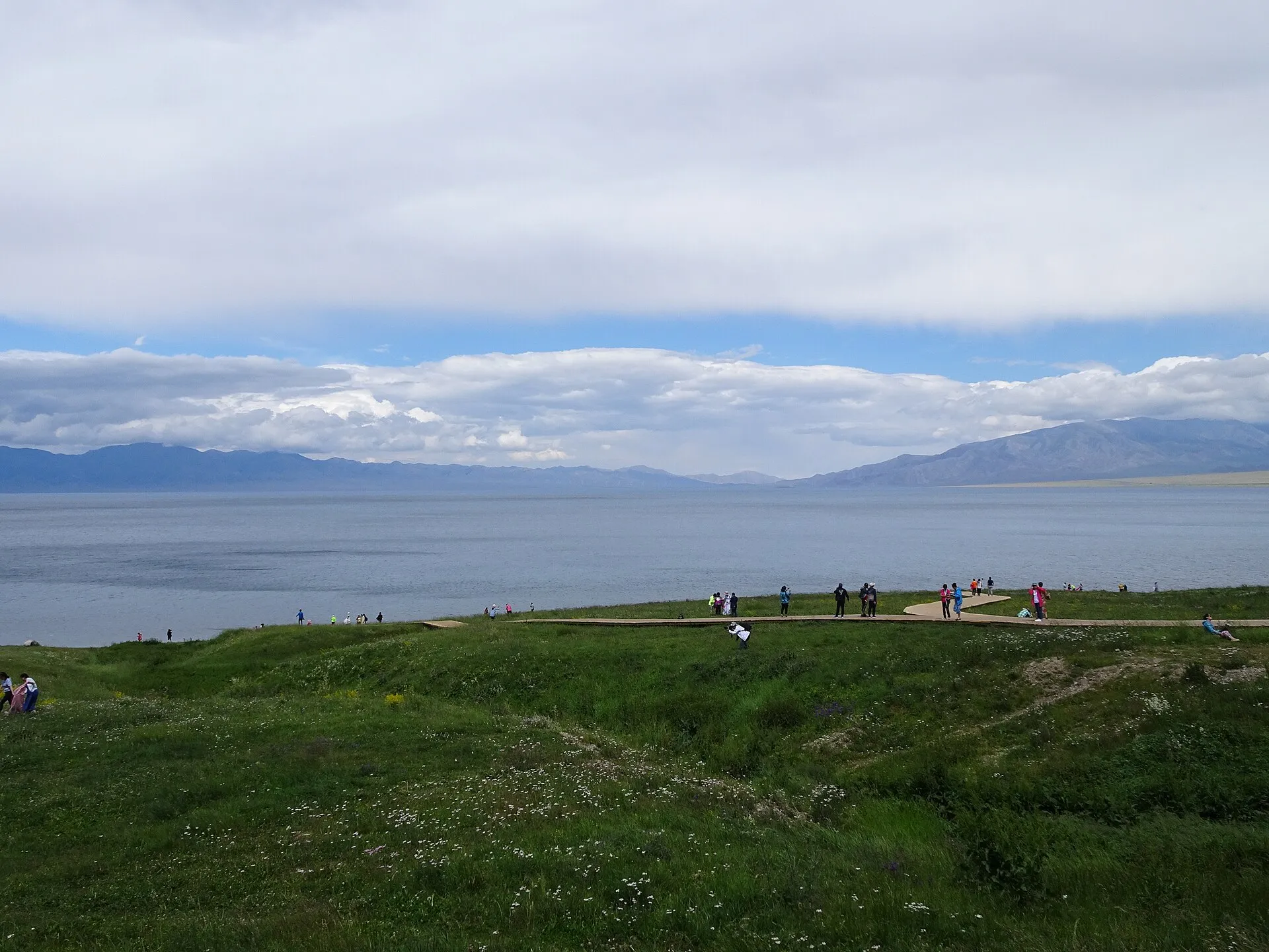 Sayram Lake, a vast blue alpine lake ringed by green grassland and distant mountains under a wide sky